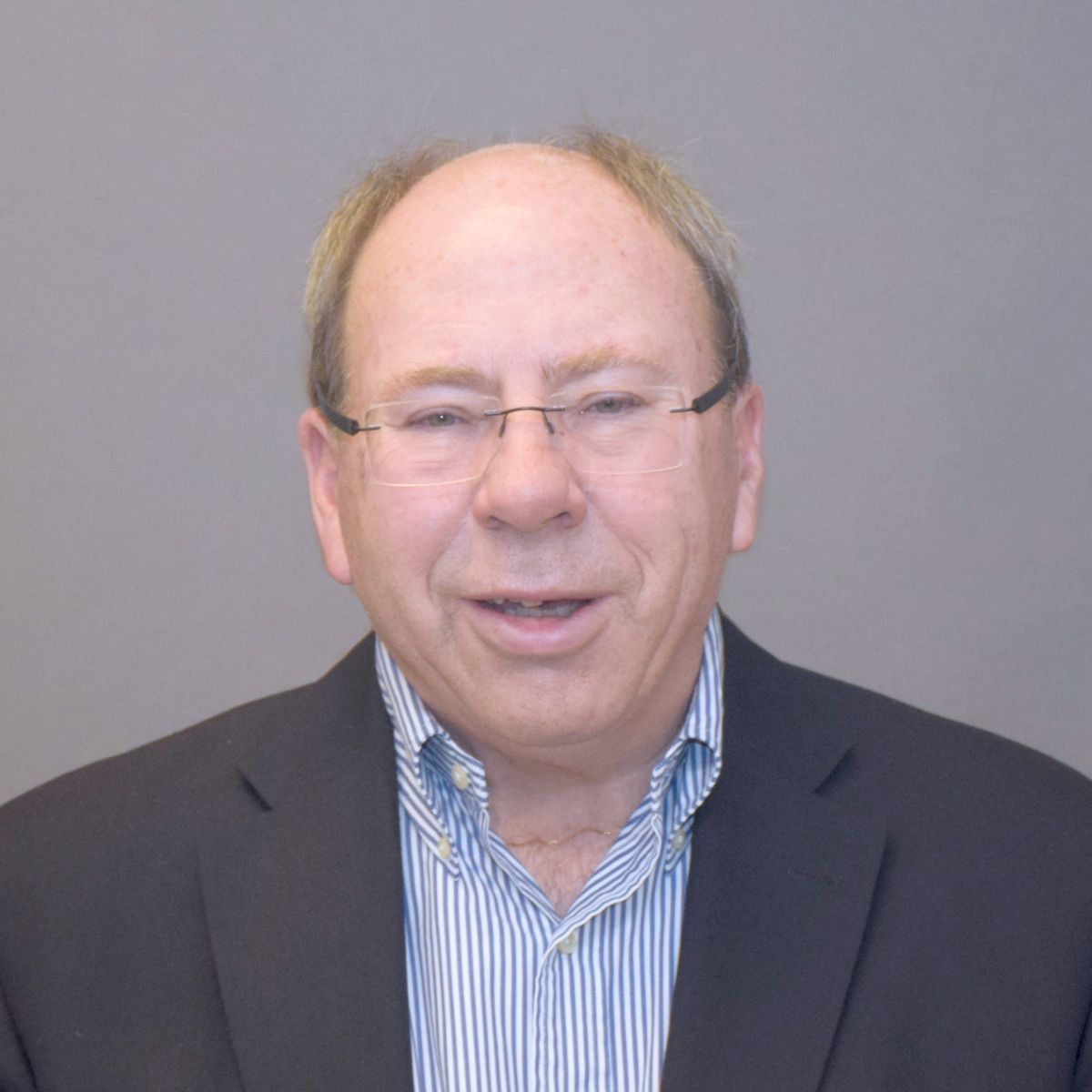 A headshot of a man wearing glasses, a blue dress shirt and a black suit jacket against a grey backdrop.