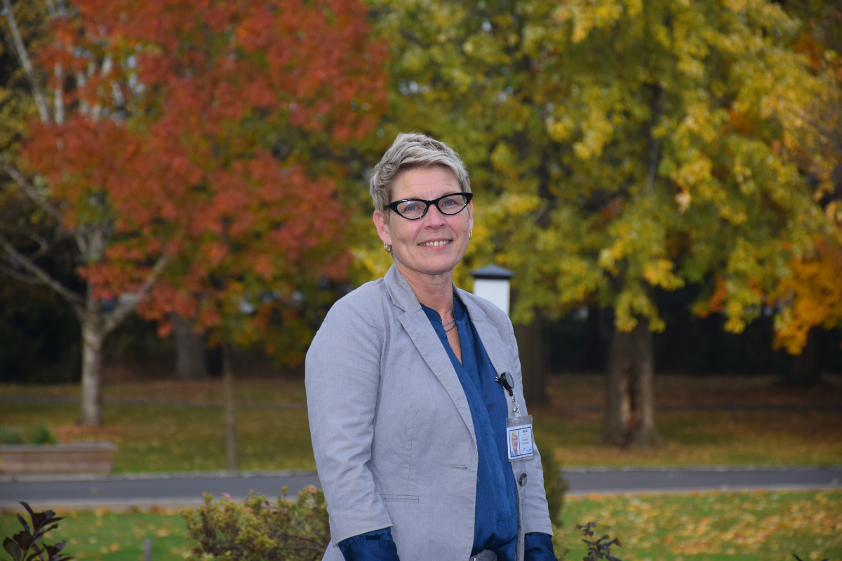 A woman with short blonde hair, black glasses and a blue shirt with a grey blazer standing outside in front of trees with fall colours.