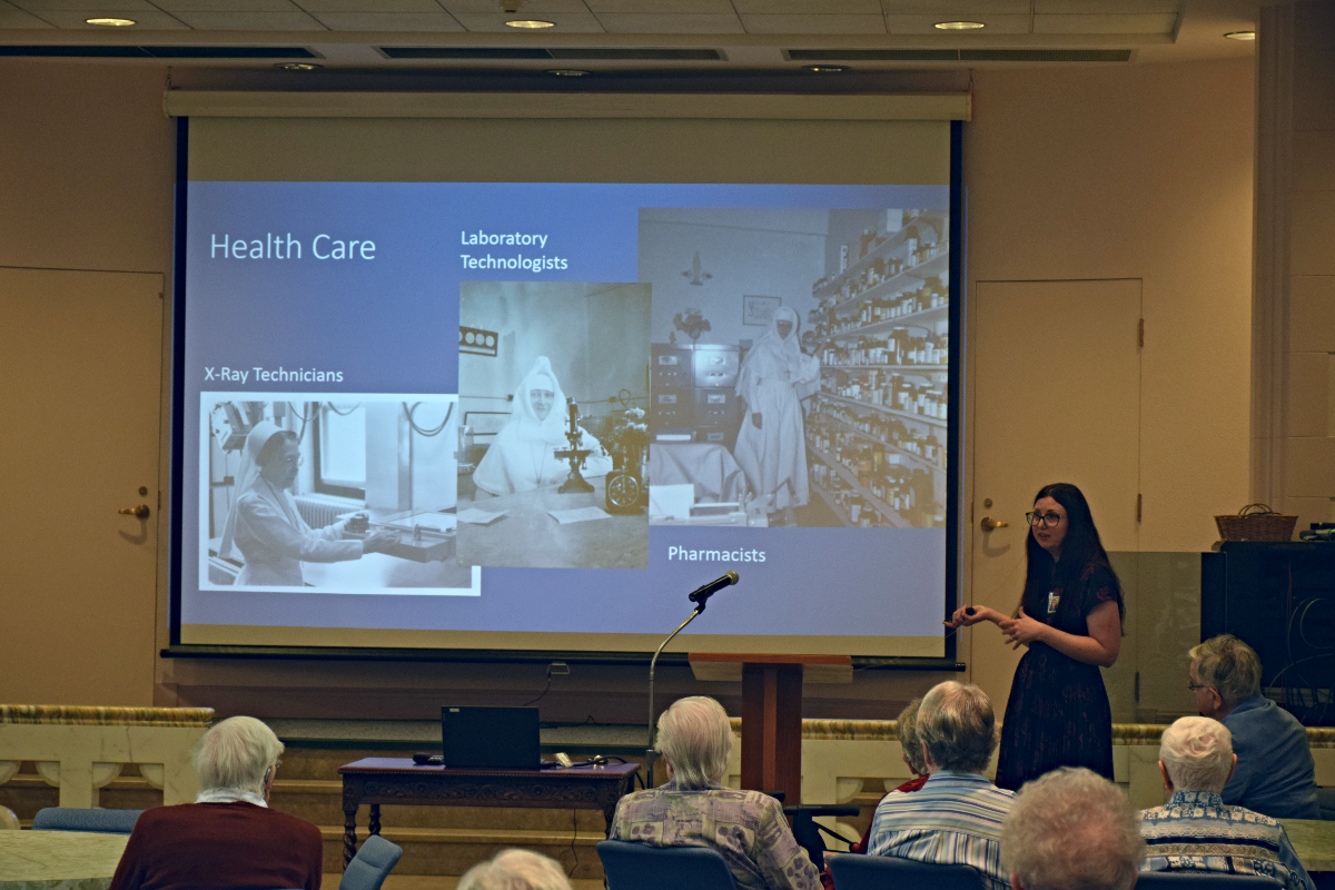 A person standing beside a projector screen in front of an auditorium full of people.