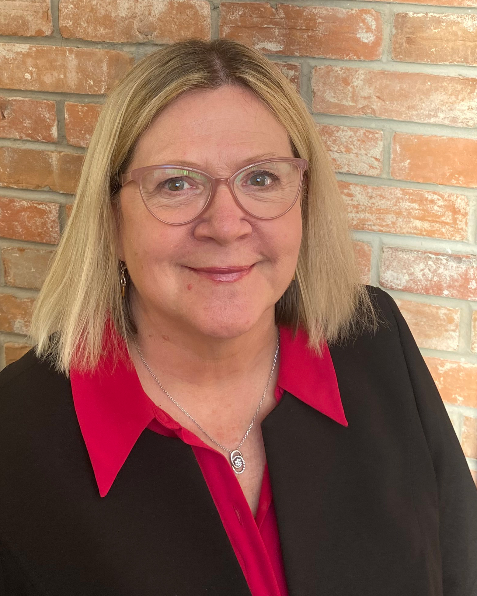Headshot of a woman wearing a red shirt, black blazer and glasses against a red brick wall.