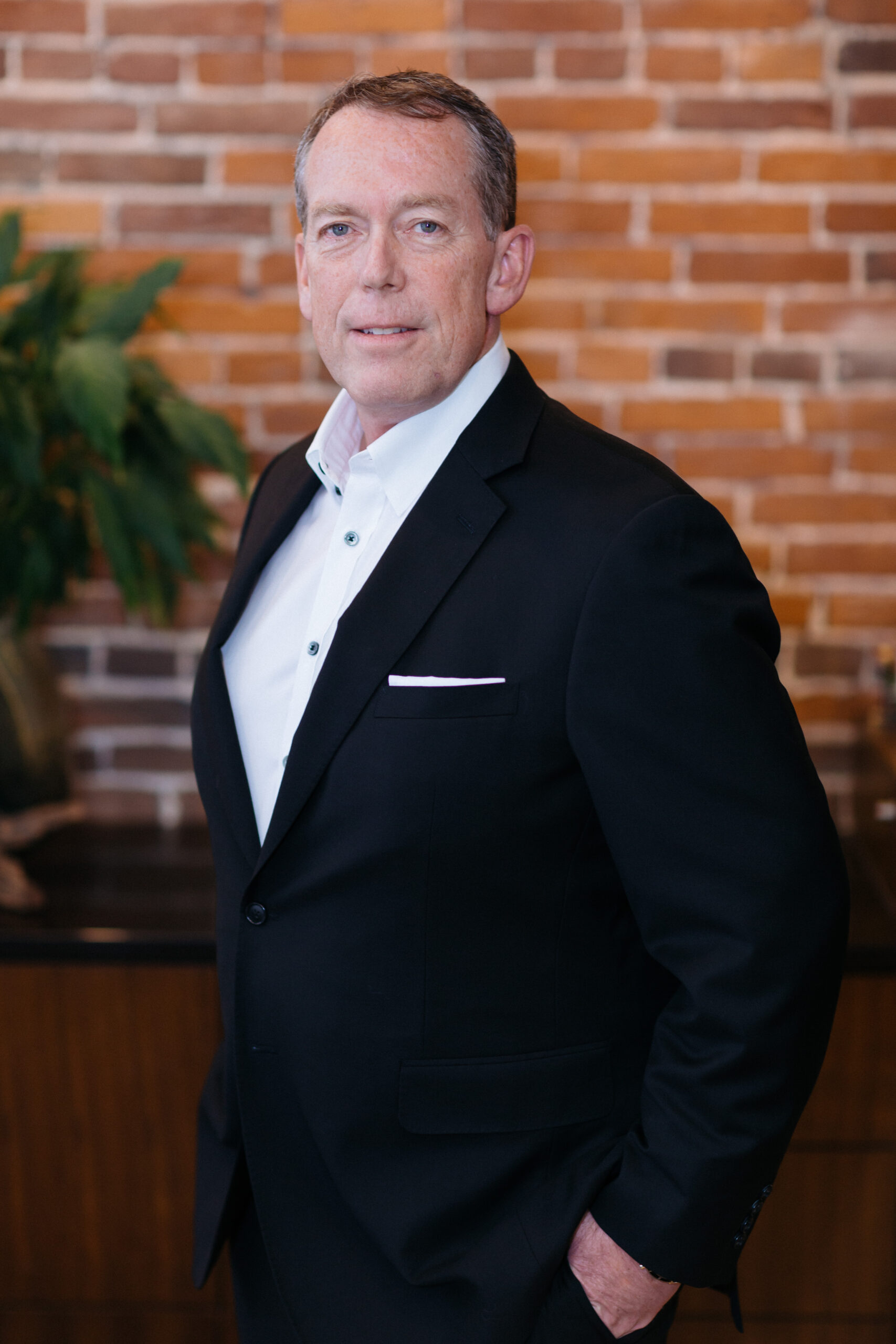 A man wearing a white dress shirt and black blazer standing in front of a red brick wall.