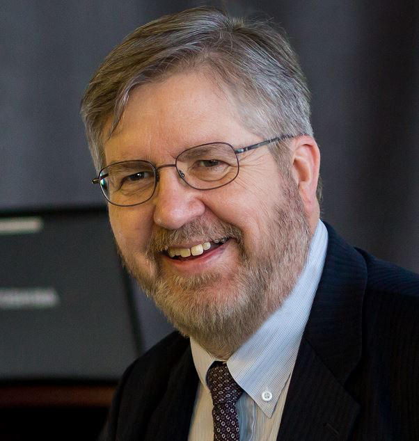 A headshot of a man wearing a white dress shirt, red tie, black suit jacket and glasses against a grey backdrop.
