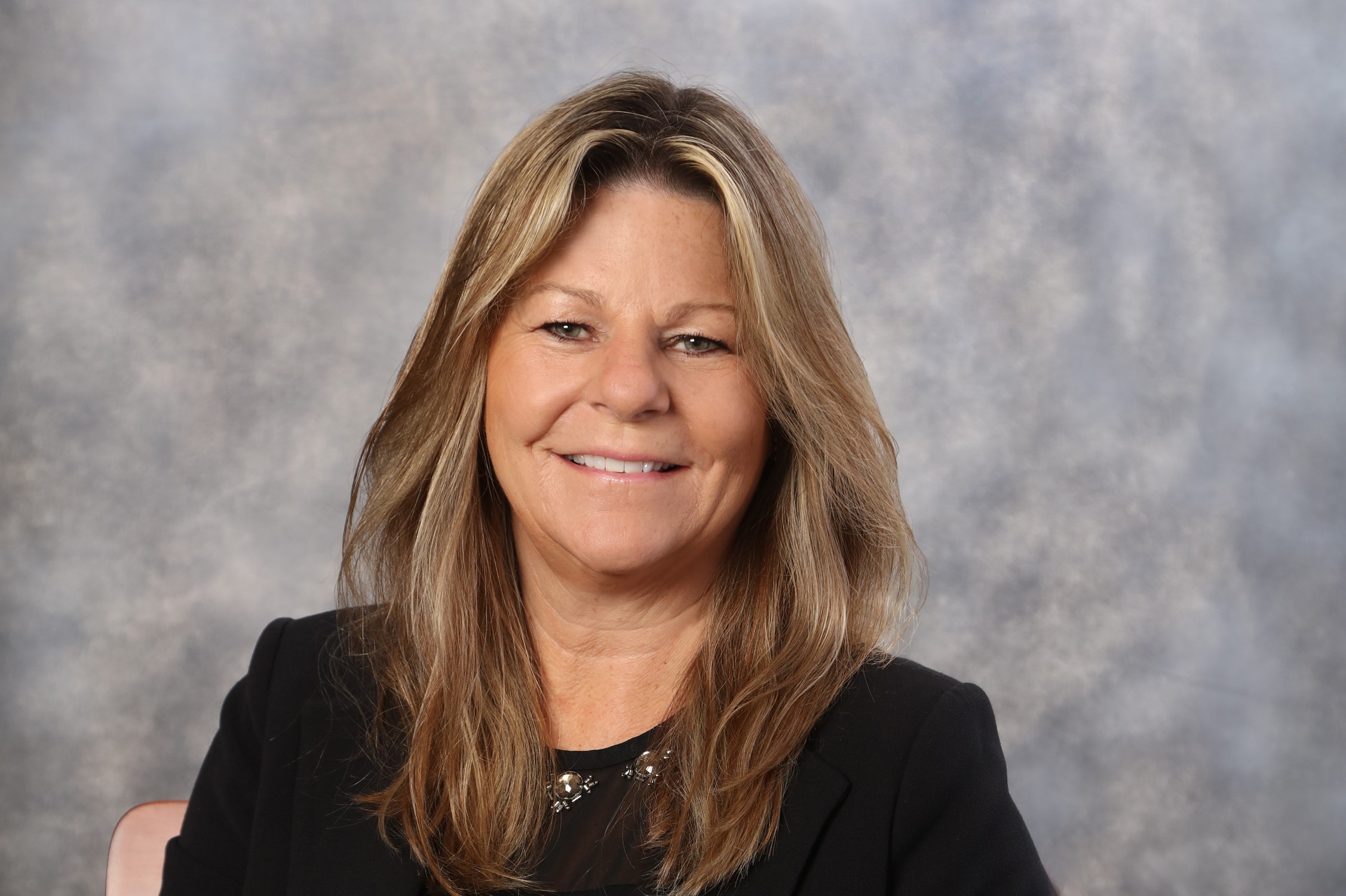 A headshot of a woman with long hair wearing a black top against a grey backdrop.