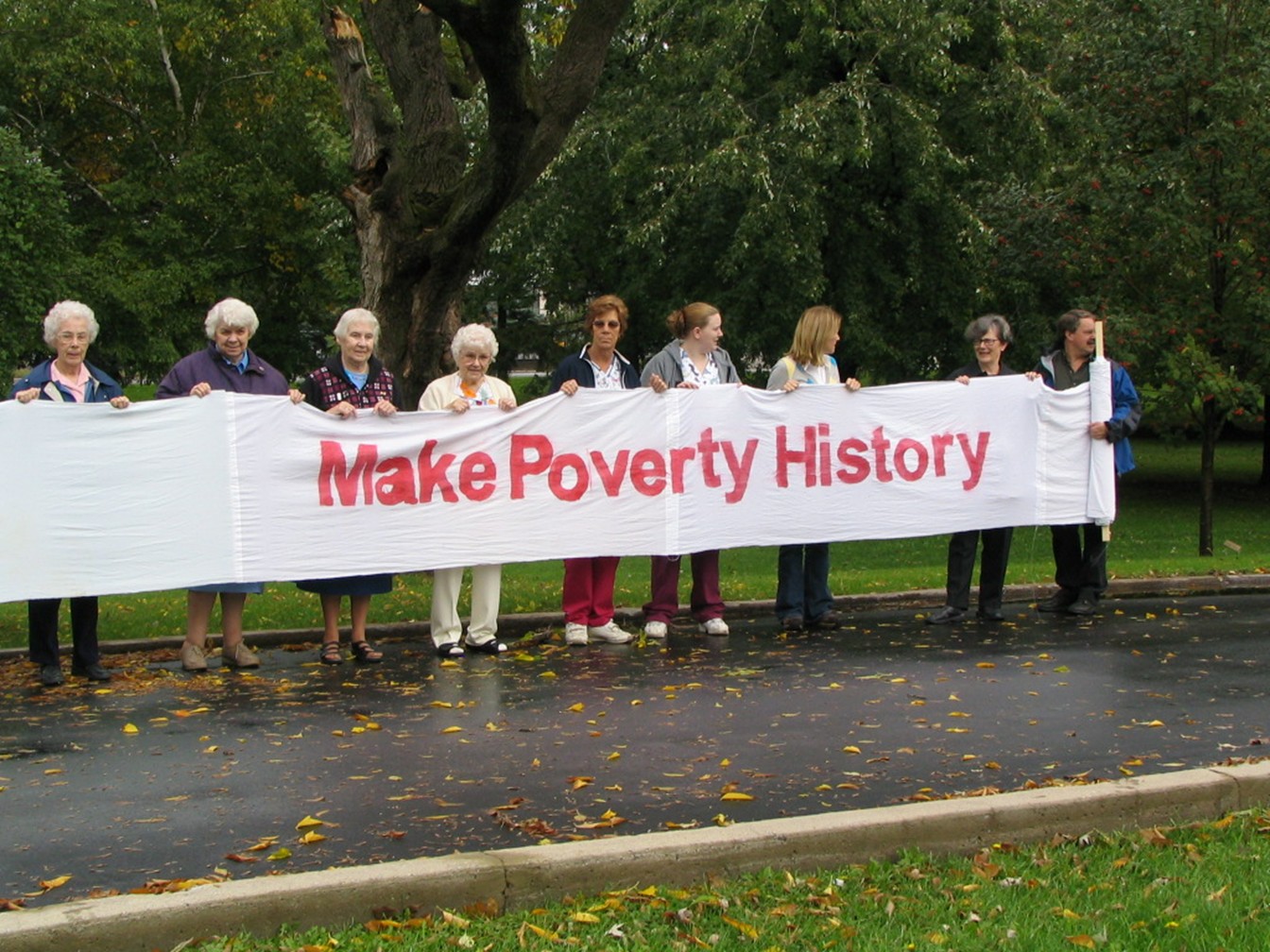 A group of Sisters of Providence and community members stand outdoors holding a large white banner that reads “Make Poverty History,” demonstrating their long-standing commitment to social justice and advocacy.