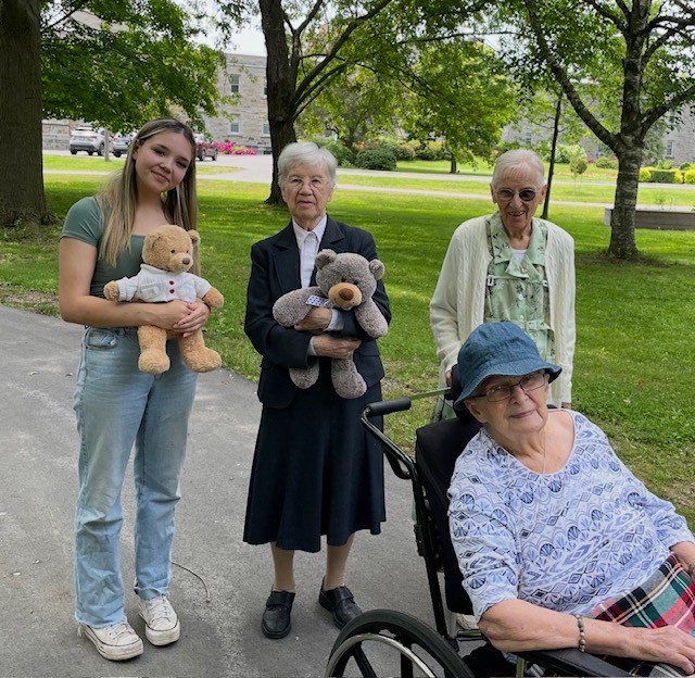 An older adult in a wheelchair, with three people standing behind her, two holding teddy bears, on a path outside with green grass and trees in the background.