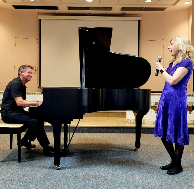 A gentleman sitting on a bench playing a black piano, looking at a woman in a blue dress singing into a microphone.