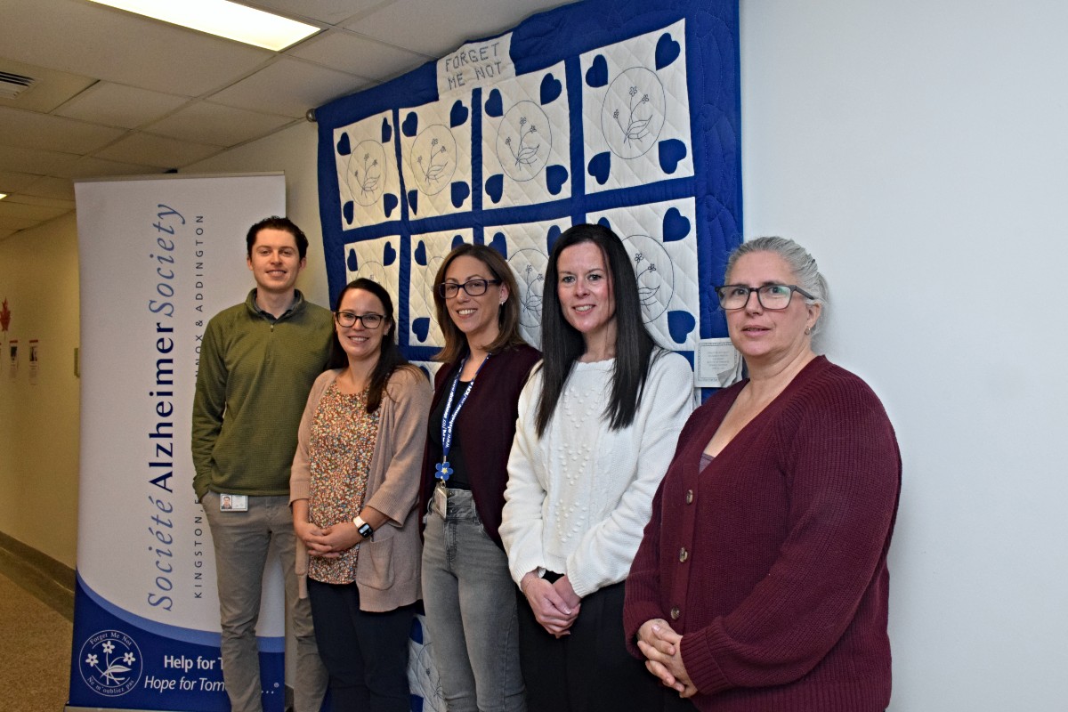 Five people stand side-by-side in a hallway in front of a blue and white quilt that is hanging on a wall.