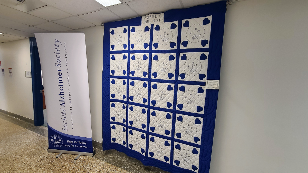 A blue and white quilt hanging on a wall, with a blue and white pull up banner displayed next to it, with the words 'The Alzheimer Society of Kingston, Frontenac, Lennox & Addington'.