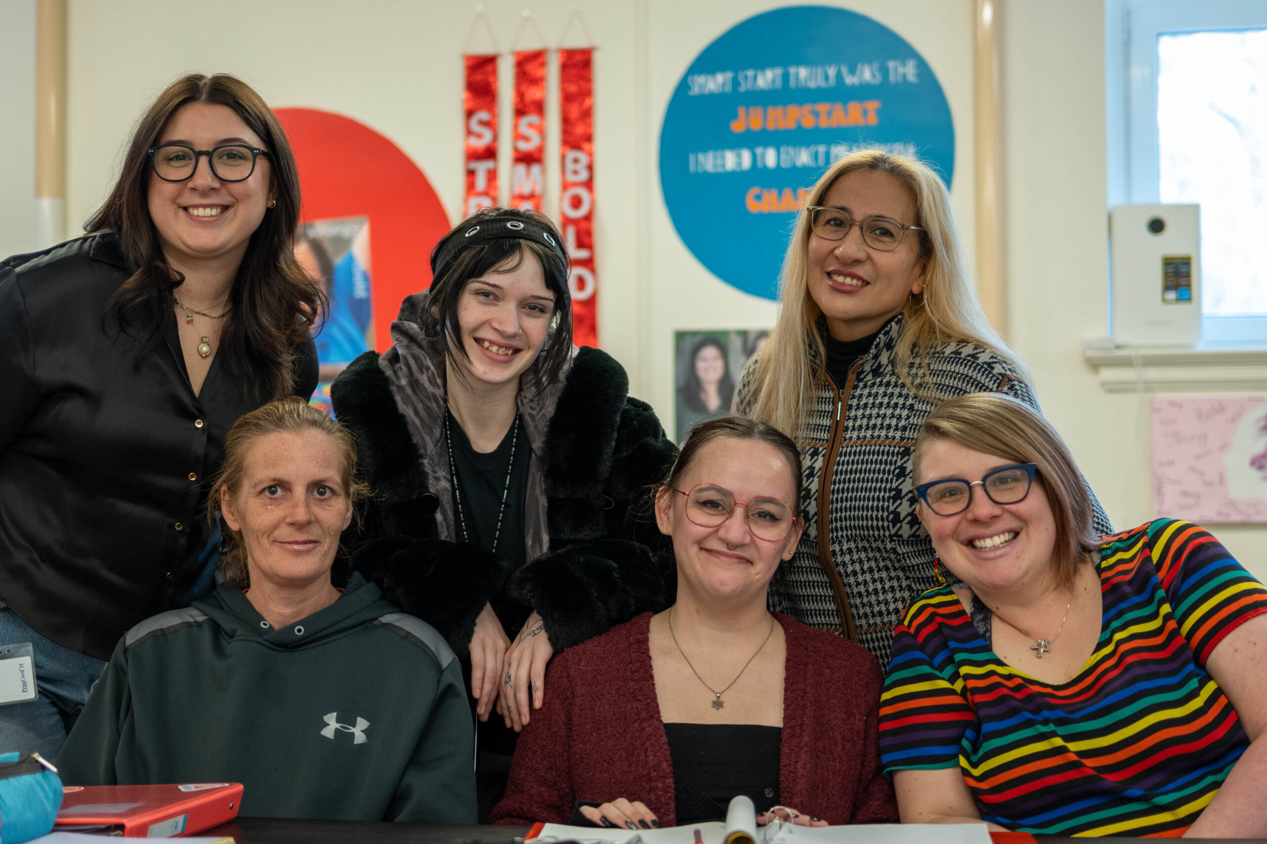 Six people grouped together, three sitting and three standing, a white wall with blue and orange posters and signs is visible behind them.