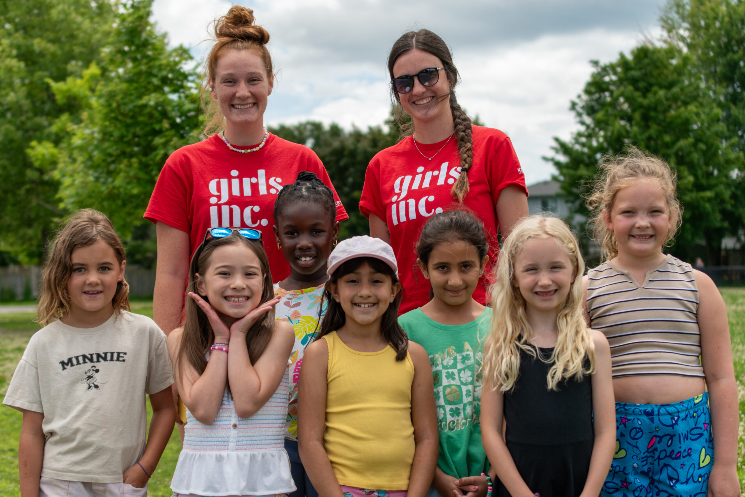 Seven young girls stand side-by-side with two adult women standing behind them outdoors in front of green trees.