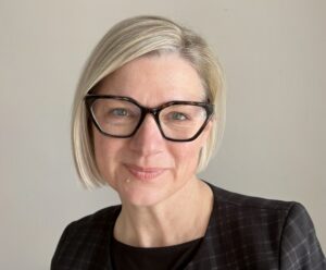 A professional headshot of a woman wearing black glasses and a black blazer.
