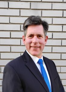 A man pictured outdoors in a suit standing in front of a light brick wall.
