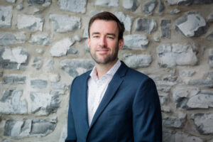 A professional headshot of a man standing against a stone wall, wearing a suit.