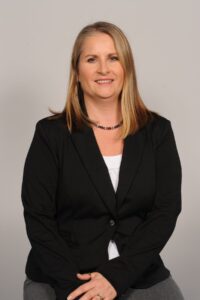 A professional headshot of a woman sitting down in front of a neutral coloured-background, wearing a black blazer.