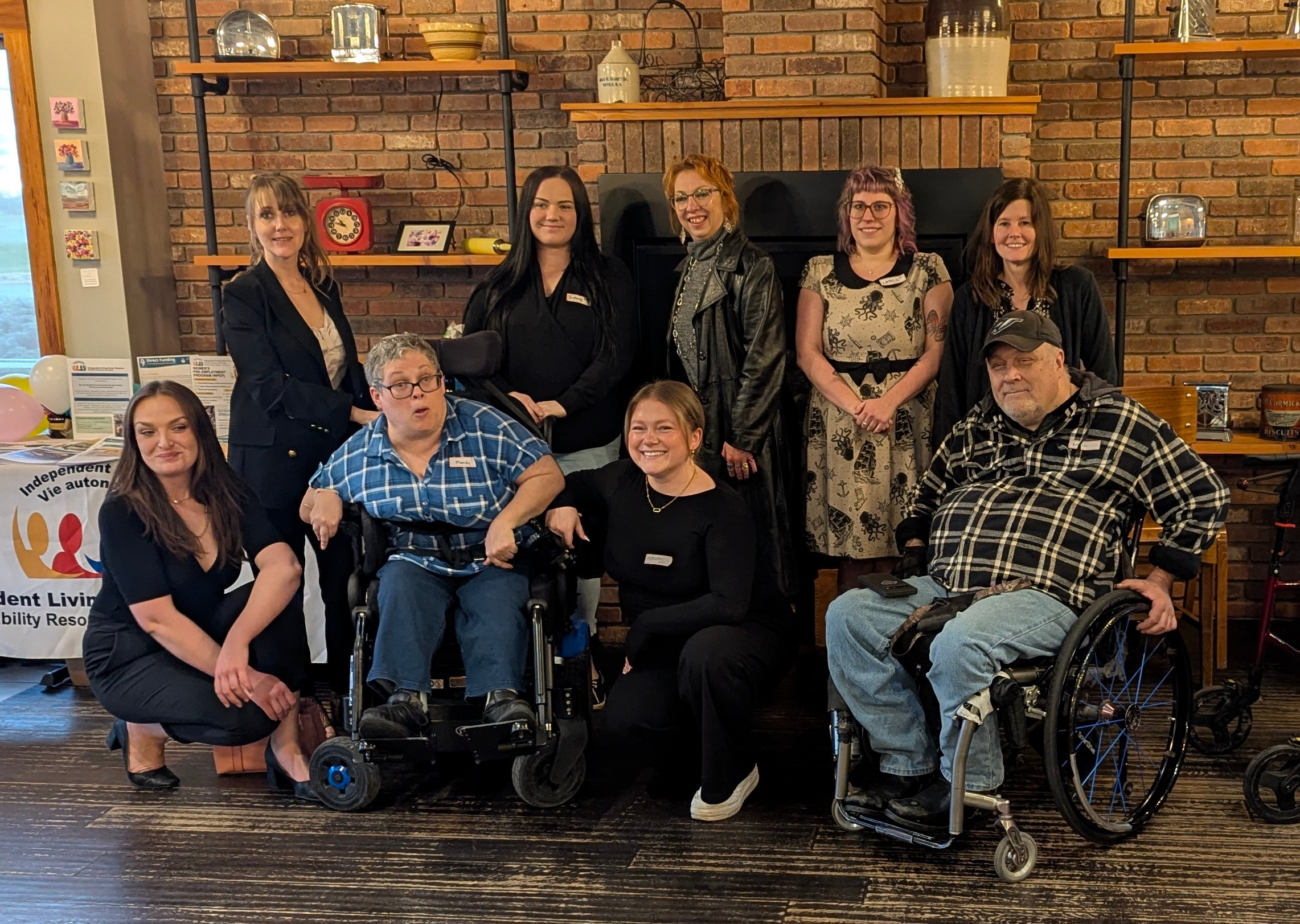 Group photo of nine people from Independent Living Centre Kingston gathered in front of a brick fireplace. Two people are seated in wheelchairs, two are kneeling in front, and five are standing behind them. An Independent Living Centre Kingston banner is partially visible on the left.