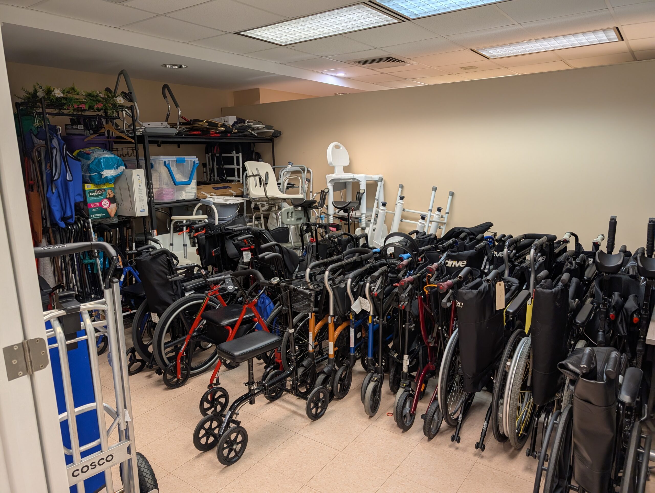 A storage room filled with mobility and accessibility equipment, including wheelchairs, walkers, rollators, canes, shower chairs and other assistive devices arranged in rows and on shelving.