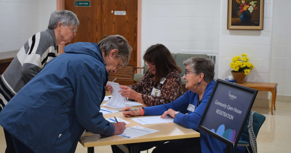 Two visitors check in at the Providence Village Community Open House while two volunteers seated behind a registration table assist them. A sign beside the table reads “Community Open House Registration.”
