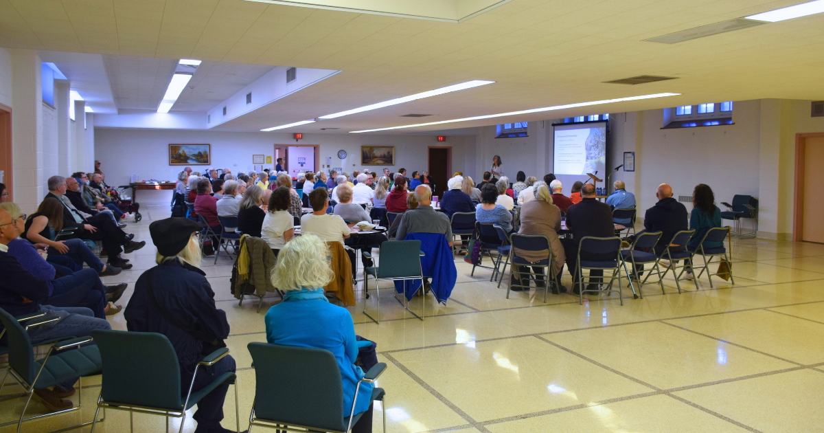 A large audience seated in rows and around tables listens to a speaker giving a presentation at the front of a bright community hall during the Providence Village Community Open House.